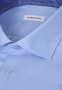 Chemise bleu clair avec col boutonné et étiquette de marque "seidensticker" visible à l'intérieur du col.