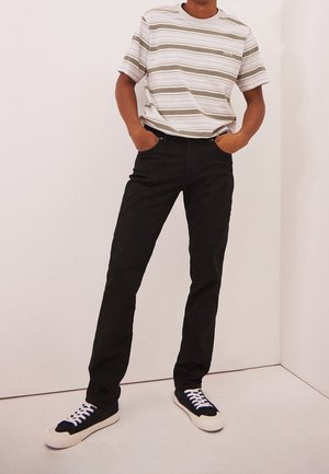 Gestreiftes T-Shirt in Weiß, Beige und Oliv, kombiniert mit schwarzen Jeans. Legere Sneakers aus schwarzem Stoff mit weißer Sohle und blauen Schnürsenkeln.