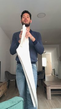 Homme debout dans un salon, tenant et pliant un tissu ou des vêtements blancs, portant un pull bleu marine et un jean bleu clair.