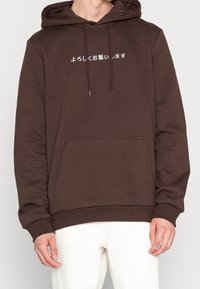 Bruine hooded sweatshirt met een voorzak en trekkoorden, met witte Japanse tekst op de borst. Zachte, textuurachtige stof.