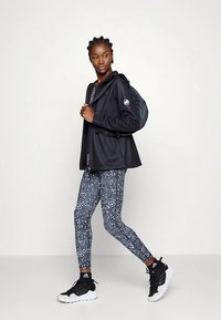 Kvinna i svart jacka, mönstrade leggings och svartvita sneakers som går med en svart ryggsäck över ena axeln.