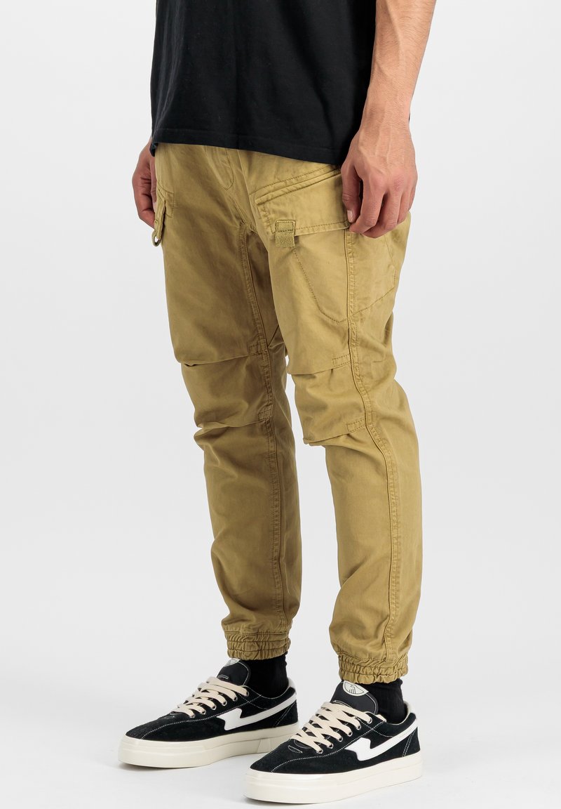 Khaki cargo pants with an elastic drawstring hem, side pockets, and relaxed fit, paired with black and white sneakers.