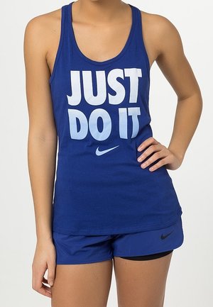 Henkilö, jolla sininen hihaton paita, jossa lukee "JUST DO IT" ja Nike-logo, yhdistettynä sinisiin Nike-shortseihin, seisoo käsi lanteilla.