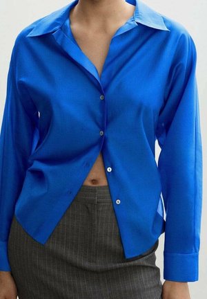 Person wearing a bright blue button-up shirt partially open, paired with high-waisted gray pinstriped trousers.