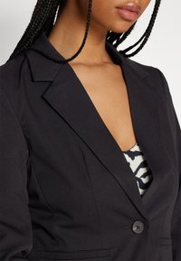 Svart kavaj med slitna kragar och enkel knappstängning, med en slät textur. Bärs över en mönstrad topp med djup u-ringning.