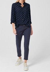 Vrouw in een marineblauwe blouse met stippen en driekwartmouwen, donkere blauwe slim fit broek en witte sneakers, staande tegen een lichte achtergrond.