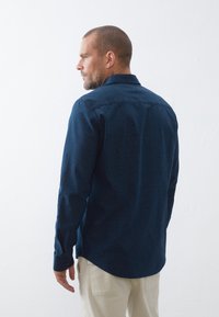 Camisa de manga larga azul marino con un pequeño patrón de puntos blancos, con cuello puntiagudo y dobladillo redondeado, hecha de una tela suave.