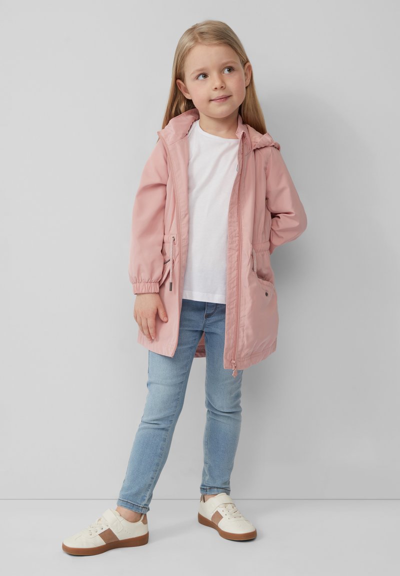 Różowa, lekka kurtka z kapturem, zapinana na zamek, z elastycznymi mankietami i kieszeniami, w połączeniu z białym t-shirtem, niebieskimi dżinsami i kremowymi trampkami z brązowymi akcentami.