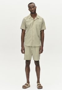 Camicia a maniche corte, di un verde chiaro, con texture e tasca, abbinata a pantaloni corti coordinati e sandali color tan, in piedi su uno sfondo semplice.