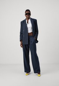 Costume à rayures bleu marine avec une veste croisée et un pantalon à jambes larges, associé à un crop top blanc et des escarpins jaunes à bout pointu.