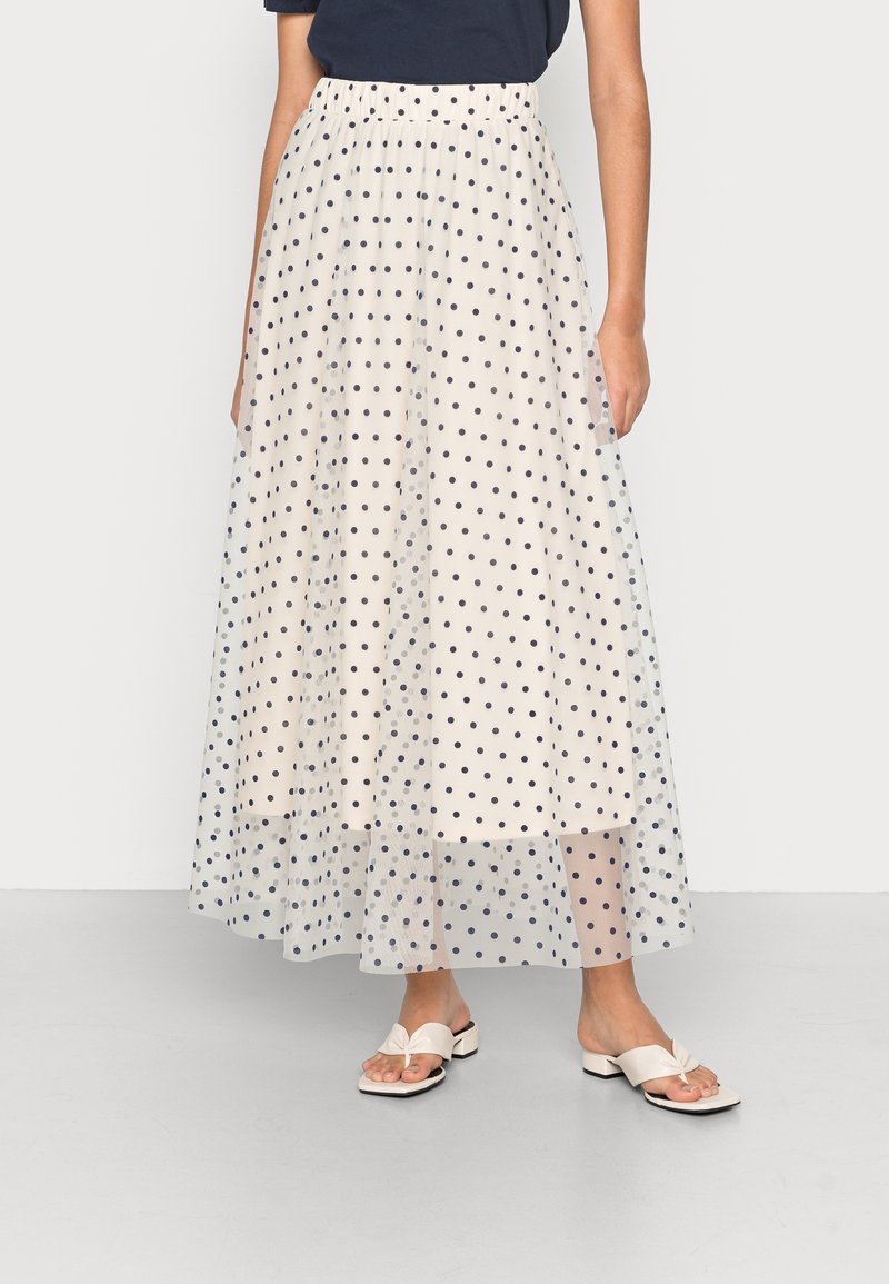 Femme portant une jupe longue blanche à pois noirs avec un voile transparent, associée à des sandales blanches ouvertes à talons bas.
