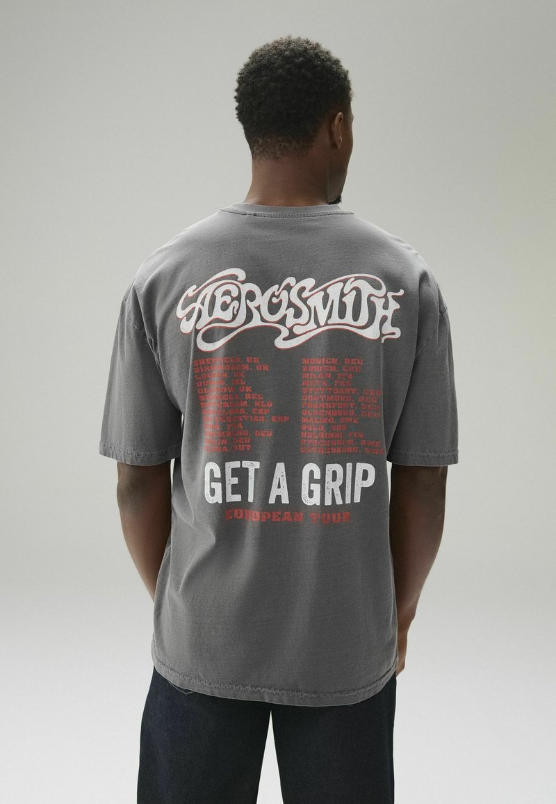 Homme portant un t-shirt gris du groupe Aerosmith avec des dates de tournée européennes en rouge et blanc et le texte "Get a Grip" au dos, debout devant un fond uni.