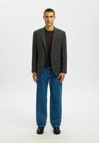 Pelēka pledēta žakete ar kontrastējošu brūnu T-kreklu un platu zilas krāsas džinsu bikses; modelis stāv melnos apavos uz neitrālas fonu.