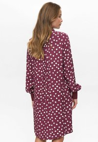 Femme aux longs cheveux portant une robe ample à manches longues de couleur bordeaux avec des pois irréguliers blancs, vue de dos sur un fond uni.
