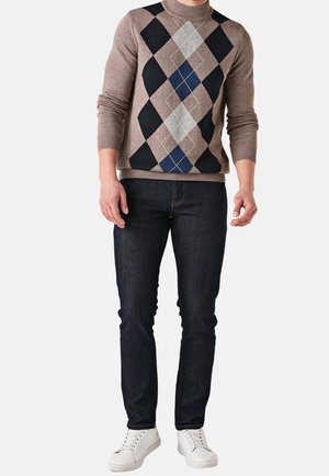 Man die een beige argyle trui draagt, donkerblauwe spijkerbroek en witte sneakers, staand tegen een effen lichte achtergrond.