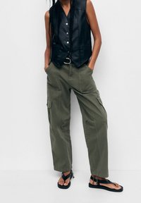 Veste noire en cuir sans manches avec boutons argentés, assortie à un pantalon cargo vert olive avec poches latérales, et sandales plates noires.