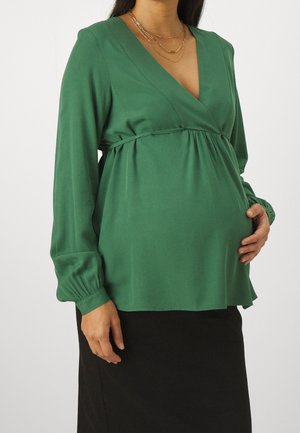 Pregnant person wearing a green long-sleeve V-neck blouse with layered gold necklaces and a black skirt, holding their belly.