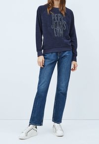 Personne portant un sweat-shirt bleu marine "Pepe Jeans", un jean droit bleu et des baskets blanches, debout devant un fond uni.