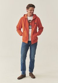 Oranje hoodie met ritssluiting, trekkoorden, steekzakken aan de voorkant en een logo patch, gecombineerd met een wit T-shirt met opdruk en blauwe jeans. Bruine laarzen.