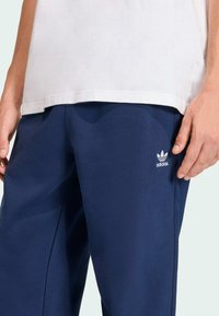 Calças de treino azul marinho com um corte descontraído. Apresentam o logótipo da Adidas bordado em branco. Feitas de um tecido suave, com uma textura lisa e detalhes mínimos.
