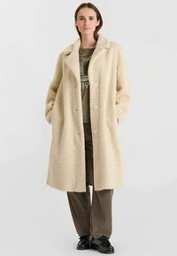 Cappotto lungo color crema, soffice, con collo con risvolto, chiusura con bottoni e tasche laterali, indossato sopra una camicia grigia e pantaloni di velluto a coste marroni.