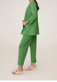 Zelený lněný oblek s volnou bundou a zúženými kalhotami. Bílé podpatečkové sandály. Jednoduchý design s knoflíkovými detaily na rukávech bundy.