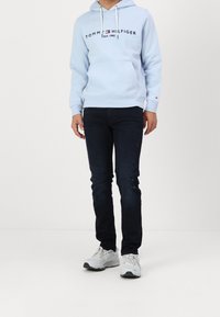 Felpa blu chiaro con logo, cordini e tasca frontale; abbinata a jeans scuri e sneaker grigie. Vestibilità casual, materiale in cotone.