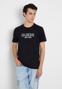 Camiseta de algodón negra con el texto "GUESS EST. 1981" en azul y gris. Cuello redondo, mangas cortas, combinada con pantalones cortos cargo en azul claro.