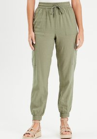 Pantalon cargo vert olive avec taille élastique, poches latérales et poignets resserrés, en tissu léger, associé à des sandales camel.