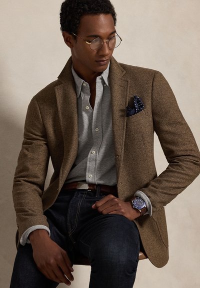 Mand iført runde briller, brun blazer med prikket lommetørklæde, grå knap-på-skjorte, mørke jeans og et armbåndsur, siddende og kiggende ned.