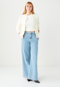 Licht crèmekleurig bomberjack met textuurpatroon, wit aangesloten topje en lichtblauwe high-waisted wijde jeans; zwarte puntige schoenen.