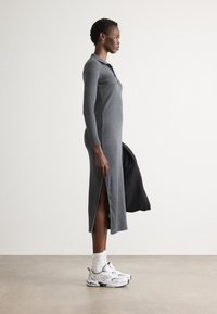 Dlhé šedé pletené šaty s gombíkovou légou a postranným rozparkom. Model má obuté biele ponožky a biele športové topánky. Hladká textúra, priliehavý strih.