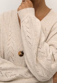 Beige pletený kardigan s hlubokým výstřihem do V, horizontální jemně žebrovanou texturou, diagonální krajkovou vzorovou aplikací a kulatým hnědým knoflíkem uprostřed.