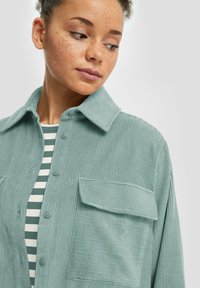 Camicia in corduroy verde chiaro con colletto alla francese, tasca anteriore e texture a righe verticali, indossata sopra un top a righe.