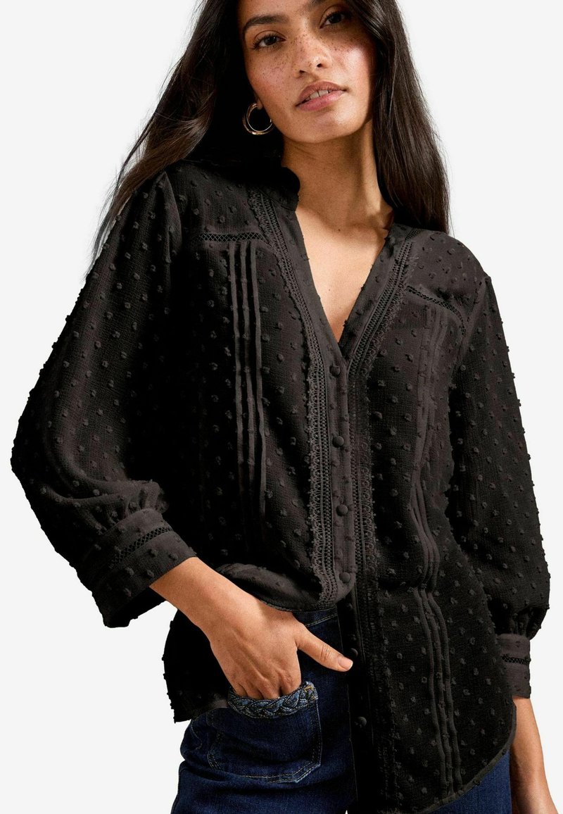 Blusa nera in tessuto testurizzato con punti in rilievo, scollatura a V, chiusura con bottoni e cuciture decorative lungo la parte anteriore e i polsini.