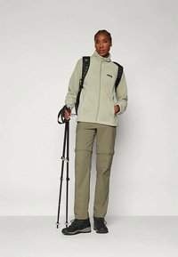 Ženska v planinski opremi drži pohodniške palice, oblečena v lahkoten flis jopič, cargo hlače, pohodne čevlje in črn nahrbtnik.