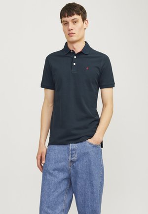 Navyblauwe polo met een kraag, drie witte knopen en een klein rood logo op de borst. Model draagt lichtblauwe denim jeans.