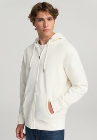 Giovane uomo che indossa una felpa bianca con zip, tasche frontali e cordini, abbinata a jeans blu, in piedi contro uno sfondo semplice.
