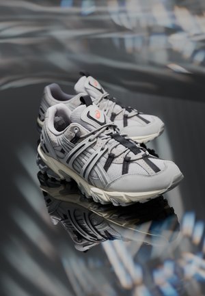 Paire de baskets athlétiques grises et noires avec semelles texturées reflétées sur une surface sombre et brillante.