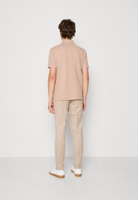 Jasno beżowa koszulka polo z krótkim rękawem, w zestawieniu z jasnobeżowymi spodniami lnianymi i białymi sneakersami, widziana z tyłu na jednolitym tle.