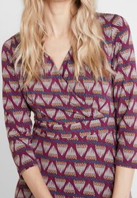 Vestido wrap de tejido morado con un patrón geométrico y acentos en crema y colores. Mangas de tres cuartos y un diseño de escote en V.
