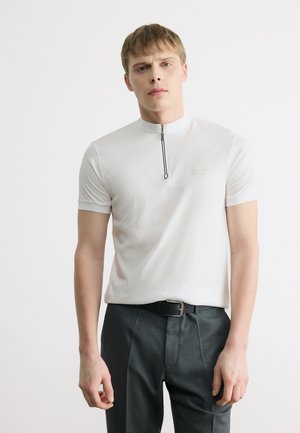 Mladý muž oblečený v bielom krátkorm rukáve so zipsom a tmavých nohaviciach, stojaci na jednoduchom bielom pozadí.