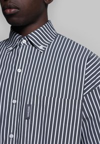 Camicia a righe grigia e bianca, dotata di colletto con bottoni, bottoni bianchi e taschino sul petto con etichetta del marchio. Tessuto textured.