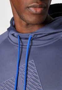 Primo piano della parte inferiore del volto e del collo di una persona che indossa un hoodie blu con lacci blu vivaci e un design geometrico testurizzato sul petto.