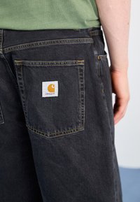Pantalones de mezclilla negros con un parche del logo de Carhartt en el bolsillo trasero. Costuras amarillas delinean los bolsillos, mostrando una tela duradera y texturizada.