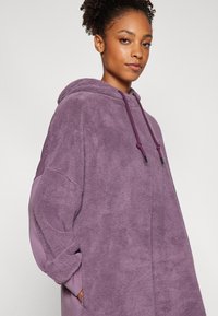 Lilla fleece hettegenser med en myk, teksturert overflate, justerbare snorer og en avslappet passform, med vide ermer og kengurulomme.