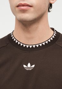 Κοντινό πλάνο του κάτω μέρους προσώπου και του λαιμού ενός ατόμου που φοράει καφέ φούτερ με λευκό λογότυπο Adidas και σχέδιο στον γιακά.