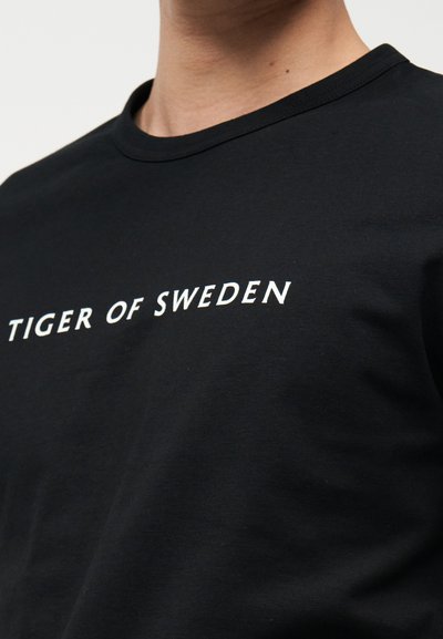 Κοντινό πλάνο ενός ατόμου που φοράει μαύρο μπλουζάκι με στρογγυλή λαιμόκοψη, με το λευκό κείμενο "TIGER OF SWEDEN" τυπωμένο στο ύψος του στήθους.