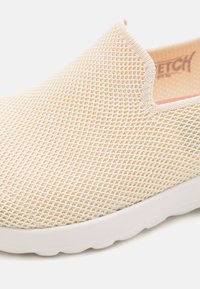 Buty wsuwane wykonane z beżowego siateczkowego materiału, z teksturowaną powierzchnią, białą gumową podeszwą i bez widocznych zapięć czy zapięć.