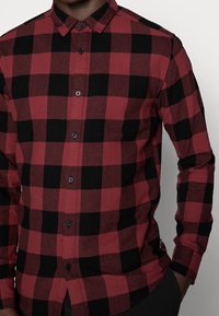 Chemise à manches longues à carreaux rouges et noirs, fabriquée dans un tissu à la texture lisse, dotée d'un col boutonné et de boutons noirs.